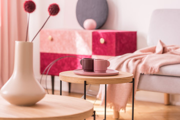 Round wooden table with cups on pink plate in living room interior with flowers in vase. Real photo