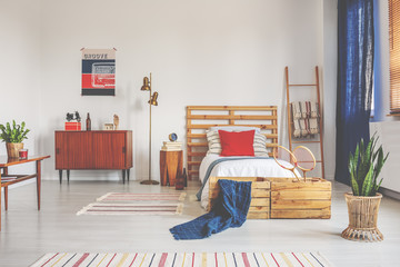 Wooden bed with white bedding and red pillow in spacious bedroom with striped carpet and retro...