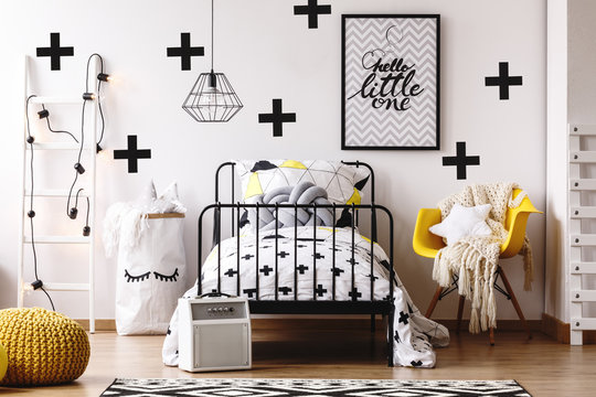 Yellow chair with blanket and pillow next to single metal bed with patterned bedding and knit pillow in elegant bedroom interior for kid