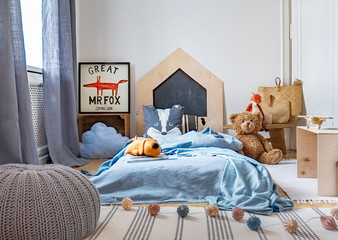 Grey pouf on carpet in child's bedroom interior with plush toy next to blue bed. Real photo
