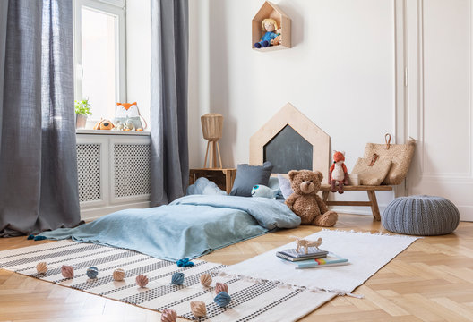 Pouf, rugs and plush toy in bright child's bedroom interior with window and blue bed. Real photo