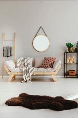 Brown fury rug on white floor of stylish ethno living room with scandinavian sofa with pillows and blanket, real photo with copy space on the empty beige wall