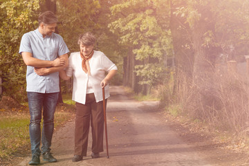 Sick grey senior lady with walking cane on the walk with handsome male nurse in the garden of nursing home - obrazy, fototapety, plakaty