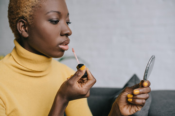 pensive african american woman applying lip gloss and looking in mirror