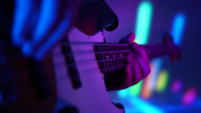 Closeup of bass player hands playing fast music with bass guitar at the concert in night club with neon lights. Modern groove music concept