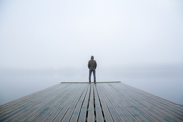 Young man standing alone on edge of footbridge and staring at lake. Mist over water. Foggy air. Early chilly morning in autumn. Beautiful freedom moment and peaceful atmosphere in nature. Back view. - obrazy, fototapety, plakaty