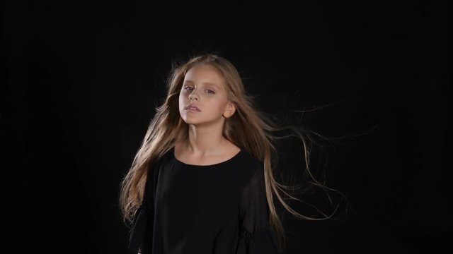 Portrait of little model in studio isolated on black background. Touching her hair in slow motion, looking at camera, posing. hd
