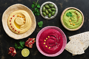 A variety of colored hummus in a plate, pomegranate, olives and pita on a dark background. Top...