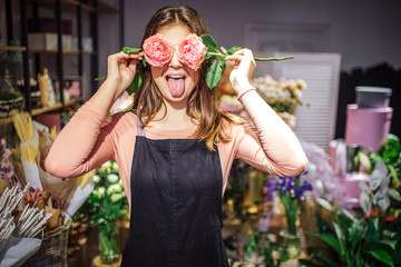 Funny and positive young female florist cover eyes with roses and show tongue. She has fun. Plants...