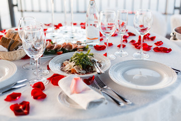 Fototapeta premium Romantic Valentine day dinner setting with rose petals and empty wineglasses on sunset
