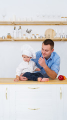 Baby boy and young father is in kitchen at table. Happy family dad, little son in the cook's cap.