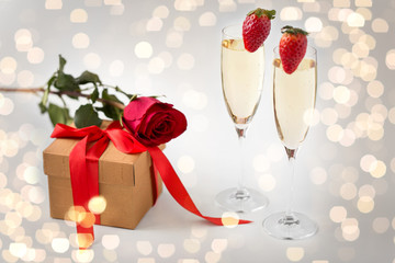 valentines day and holidays concept - two champagne glasses with strawberries and gift box with red rose over festive lights