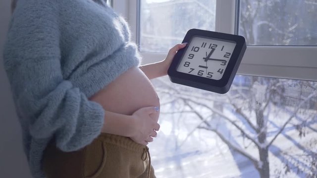 expectant mother at home holds clock in hands and stroking her naked tummy while standing by a large window