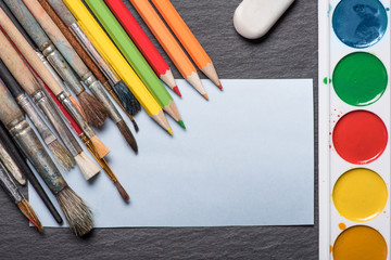 Paper, watercolors, paint brush and some art stuff on the wooden table.