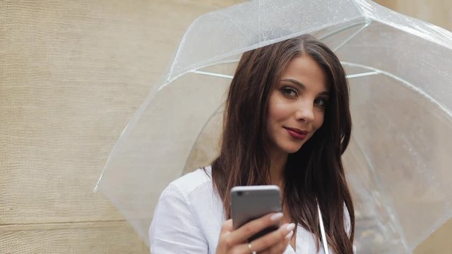 Beautiful young business woman using smartphone on the street in rainy weather, smiling, holding umbrella. She looking into the camera. Communication concept