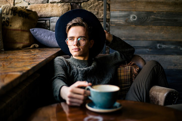 Young handsome guy in a black hat and glasses drinking coffee in a cozy cafe