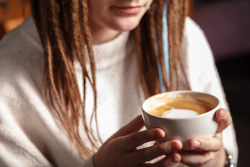 A white Cup of hot latte art coffee with a heart shape in the hands of a young girl with dreadlocks.  Woman wearing warm winter knitted white sweater. Toned. Coffee Girl Concept Relaxation Coffee Mug.