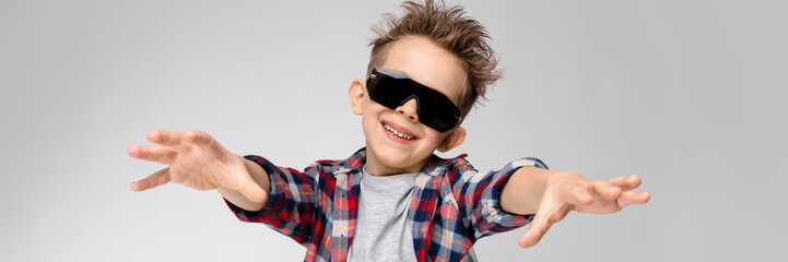 A handsome boy in a plaid shirt, gray shirt and jeans stands on a gray background. The boy in black sunglasses. The boy stretches his arms forward.