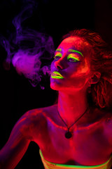 Fototapeta na wymiar Portrait of a young Smoking sexy girl with ultraviolet paint on her body. Pretty woman with glowing bodyart in black lamp light
