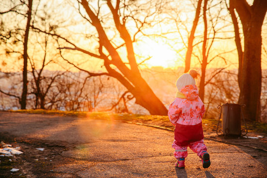 Young Little Toddler Girl Walking By City Park On Sunset. View From Behind