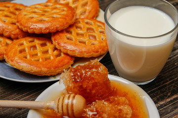 Honey with honeycomb in a white ceramic plate, cookies with jam and cup of milk on rustic wooden table