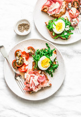 Delicious breakfast, snack, appetizers for wine, tapas - sandwiches with prosciutto, egg, arugula, cherry tomatoes and cream cheese on a light background, top view. Flat lay