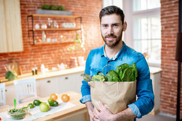 Fototapeta premium Handsome man in blue shirt standing with shopping bag full of healthy green food on the kitchen