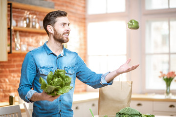 Fototapeta premium Handsome man throwing up green pepper while cooking vegan meals on the kitchen at home