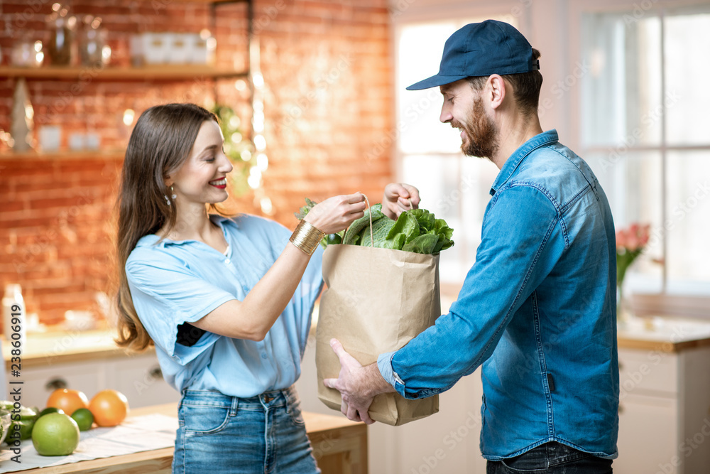 Wall mural Courier service worker delivering fresh food, giving shopping bag to a happy woman client on the kitchen at home