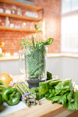 Blender full of ingredients for smoothie making on the table with green raw vegetables on the kitchen