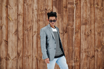 Casual young indian man in silver blazer and sunglasses posed against wooden background.