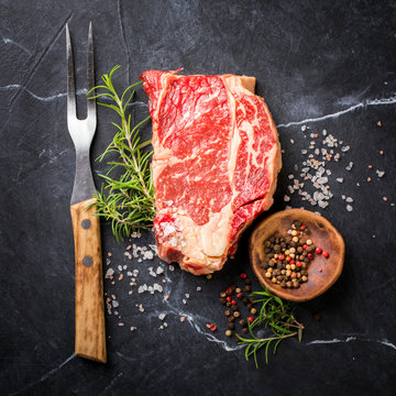 Raw Fresh Marbled Meat Beef Steak and Meat Fork Herbs and Seasonings on a black marble  Background and a cutting board Rosemary Pepper and Salt Ingredients for Cooking Top View