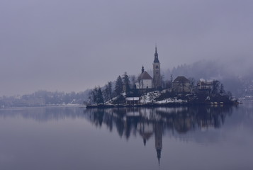 Fototapeta na wymiar Little Island with Catholic Church in Bled Lake. With reflection of church in the lake. In winter in the foggy, rainy and cloudy morning. Slovenia, Bled, February 2018