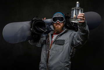 Fototapeta na wymiar Cheerful redhead man wearing a full equipment for extreme snowboarding holding a snowboard on his shoulder and celebrate victory with a bottle of alcohol in his hand.