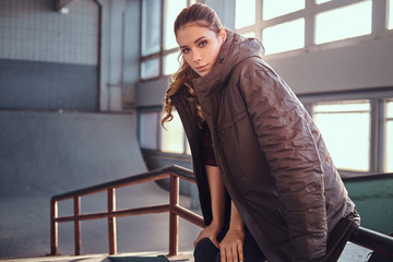 Fototapeta premium Portrait of a charming young girl with a coat covering his shoulders looking at camera while sitting on a grind rail in skatepark indoors.