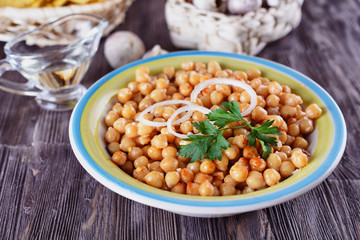 Boiled chickpeas, seasoned with vegetable oil, ground red pepper, sesame seeds and onion rings in a bowl with old spoon on a wooden background next to garlic 