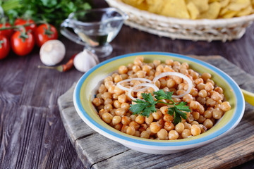 Boiled chickpeas, seasoned with vegetable oil, ground red pepper, sesame seeds and onion rings in a bowl with old spoon on a wooden background next to garlic and tomatos and corn chips