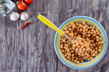 Boiled chickpeas, seasoned with vegetable oil, ground red pepper,  sesame seeds and onion rings in a bowl with old spoon on a wooden background next to garlic and tomatos, top view