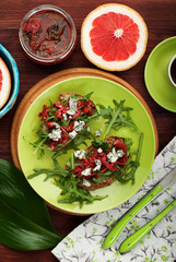 Light breakfast in a Mediterranean style. Sandwiches with rye bread, arugula and sun dried tomatoes on a green plate. Near the grapefruit and a fragment of a cup of coffee. Brown wooden background