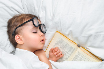 A cute toddler boy is sleeping in a round glasses with an old book under a white blanket.