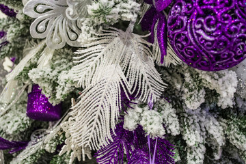 White feather and purple toys hanging on Christmas tree, covered with fake snow.