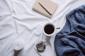 Cup coffee , blue knitted sweater, notebook with pen,  clock, candle on bed, top view. Women clothing. Flat lay, cozy concept