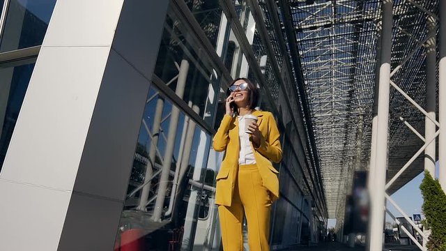Beautiful business woman in suit goes near modern business centre in the urban street, drinking coffee, communicates via smartphone. Concept of technology, lifestyle, communication people, business