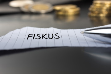 Fiskus auf einem Zettel mit Stift vor Geldmünzen