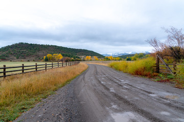 A dirt road lined by a wooden fence curves through fall colored fields