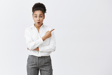 Woman being impressed and surprised coworker got promoted first. Attractive and young african-american female in pants and shirt, pointing at upper right corner, saying wow with folded lips