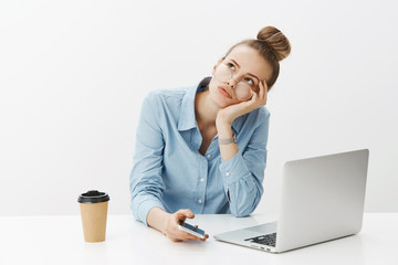 Girl dreams clock move faster as feeling bored in office, leaning head on palm gazing at upper left corner indifferent sitting near laptop and holding smartphone, daydreaming, drinking coffee