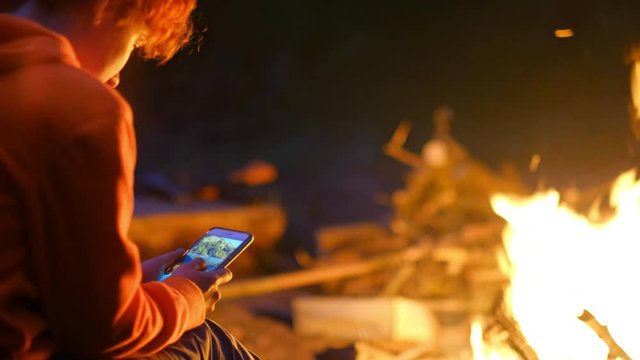 Tourist woman sitting near campfire and makes photo on smartphone at night. Bonfire fire on wild nature. Mobile photography, traveler girl and bright flame light. Cozy evening in forest outdoors.