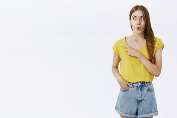 Wow looks cool. Amused and excited attractive caucasian female in yellow t-shirt folding lips from amazement and surprise gazing and pointing left with curious and interested look over white wall