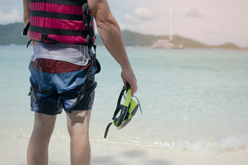 The man in  wet outfit  standing facing to the sea, holding snorkeling mask after finished snorkeling in the island